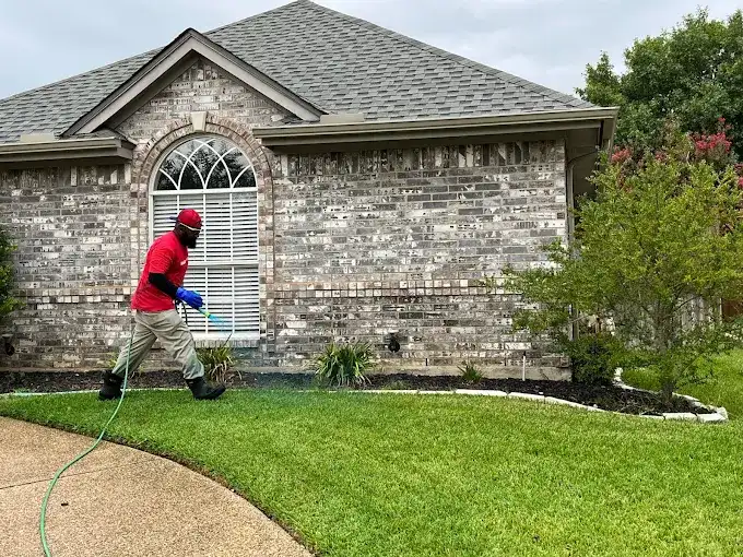 Buffalo Outdoor technician in a branded shirt spraying a perimeter barrier along the foundation of a North Texas brick home