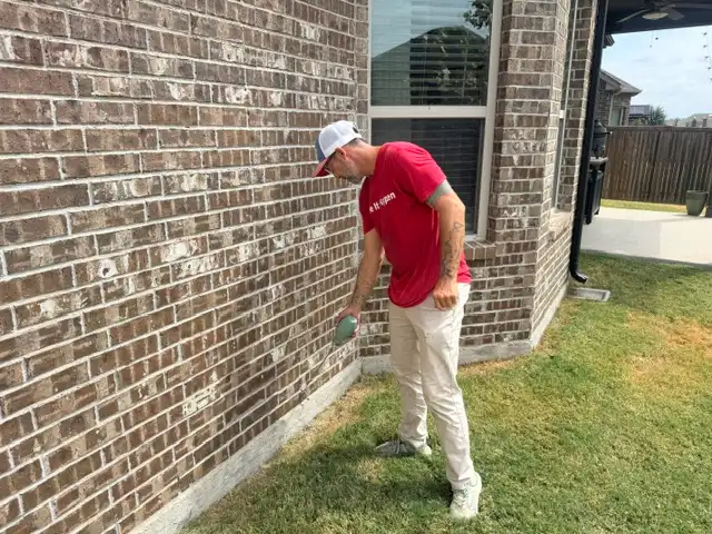 Technician dusting brick weep holes at the foundation line of a Fort Worth home